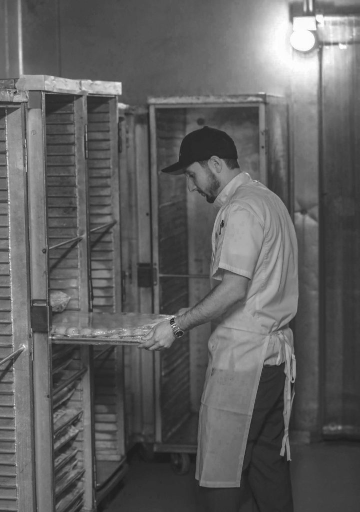 Baker carefully sliding a tray of goods into an industrial oven.