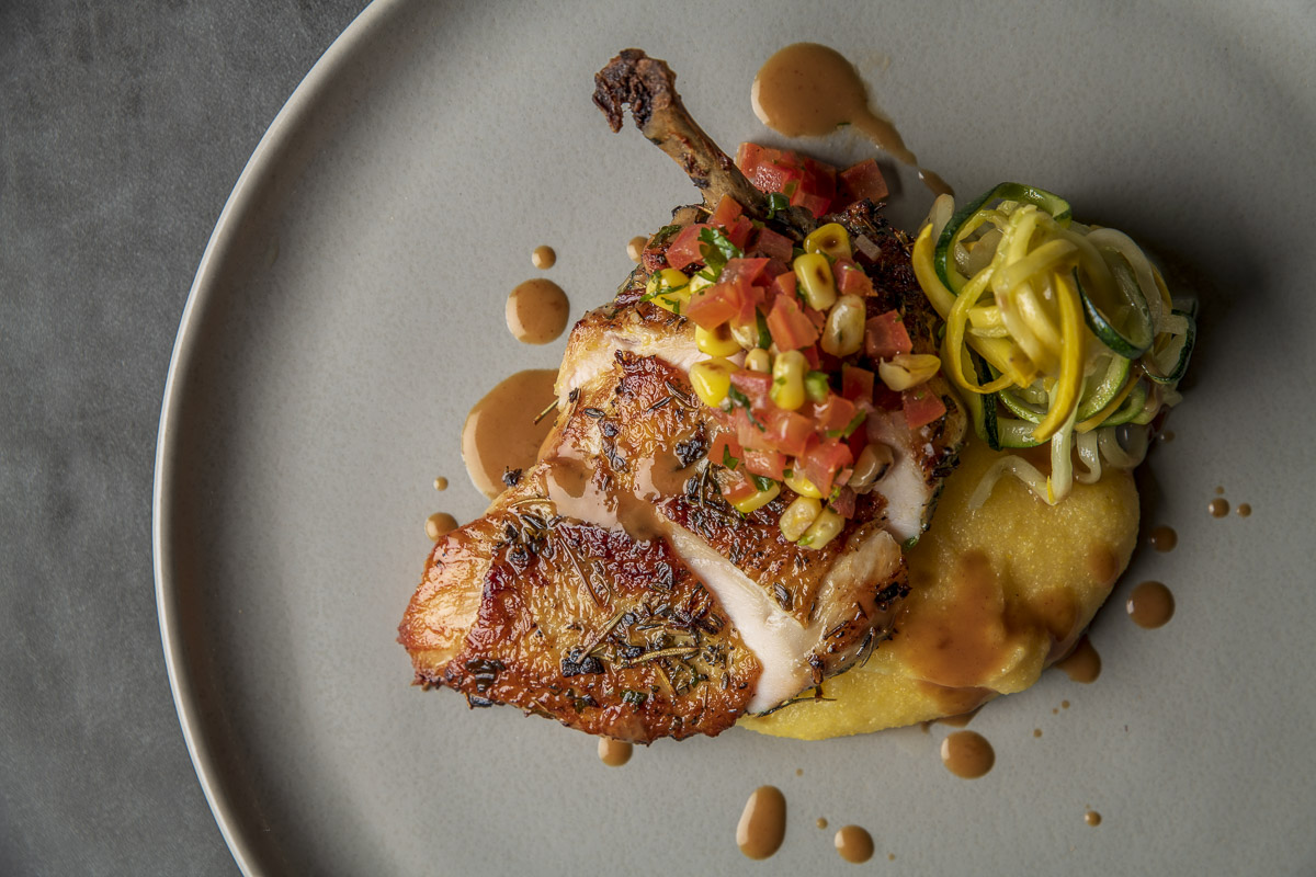 Roasted chicken thigh with a corn salsa, zucchini noodles, and a smooth puree, artistically plated on a grey dish.