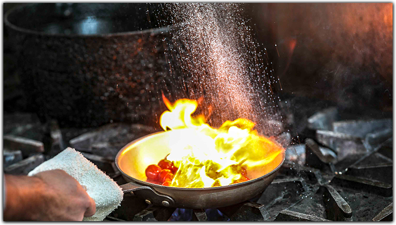 A chef flambeing food in a skillet over an open flame, with a splash of seasoning being added.