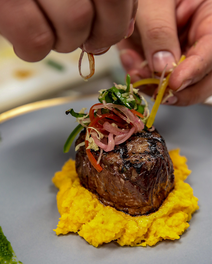 Chef garnishing a grilled steak served on a bed of mashed yellow vegetables with fresh herbs and vegetables.