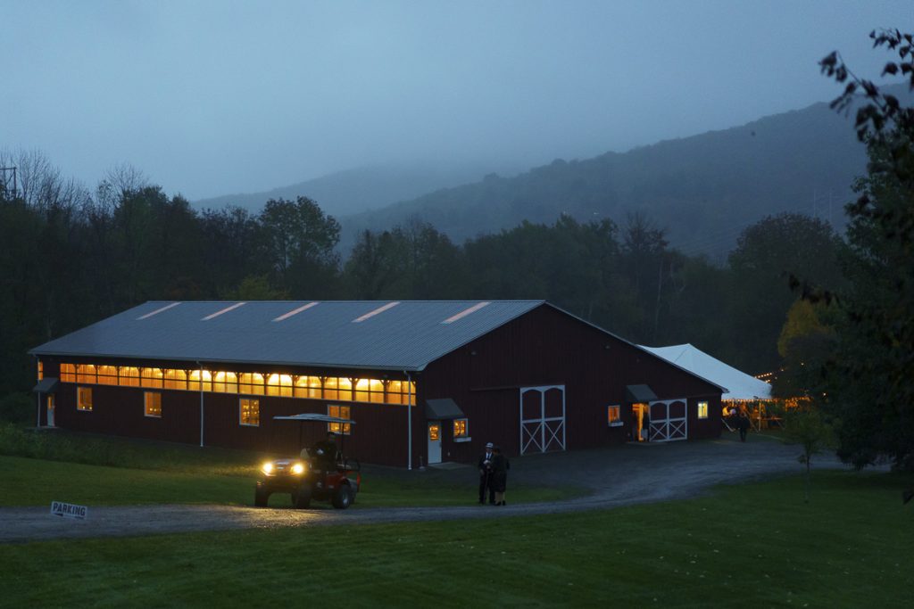 A well-lit barn at twilight with gentle fog rolling over the hills, while a vehicle approaches and two individuals stand nearby.