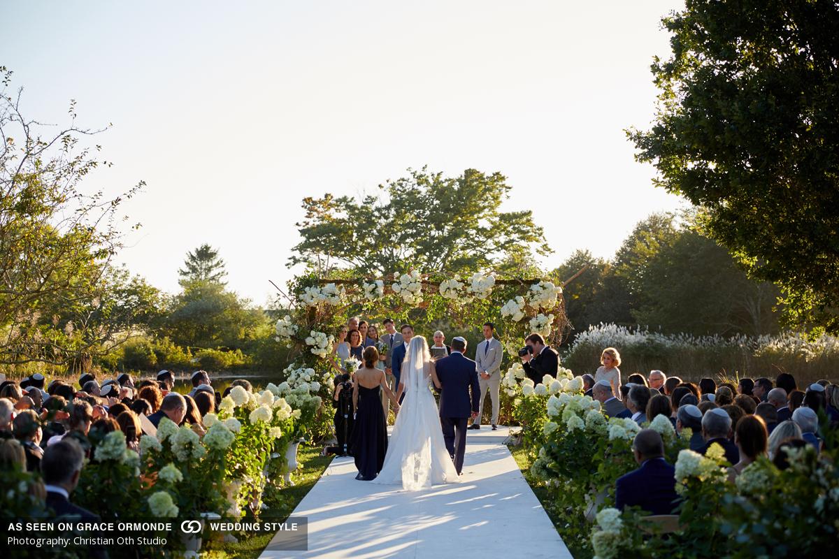 Outdoor wedding ceremony with guests seated on either side of a white aisle leading to a flower-adorned altar, with attendants standing around the couple under a clear sky.